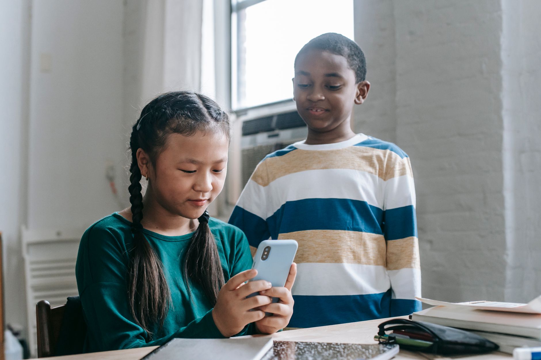 Happy little African American boy standing near Asian classmate girl using smartphone sitting at desk during break
