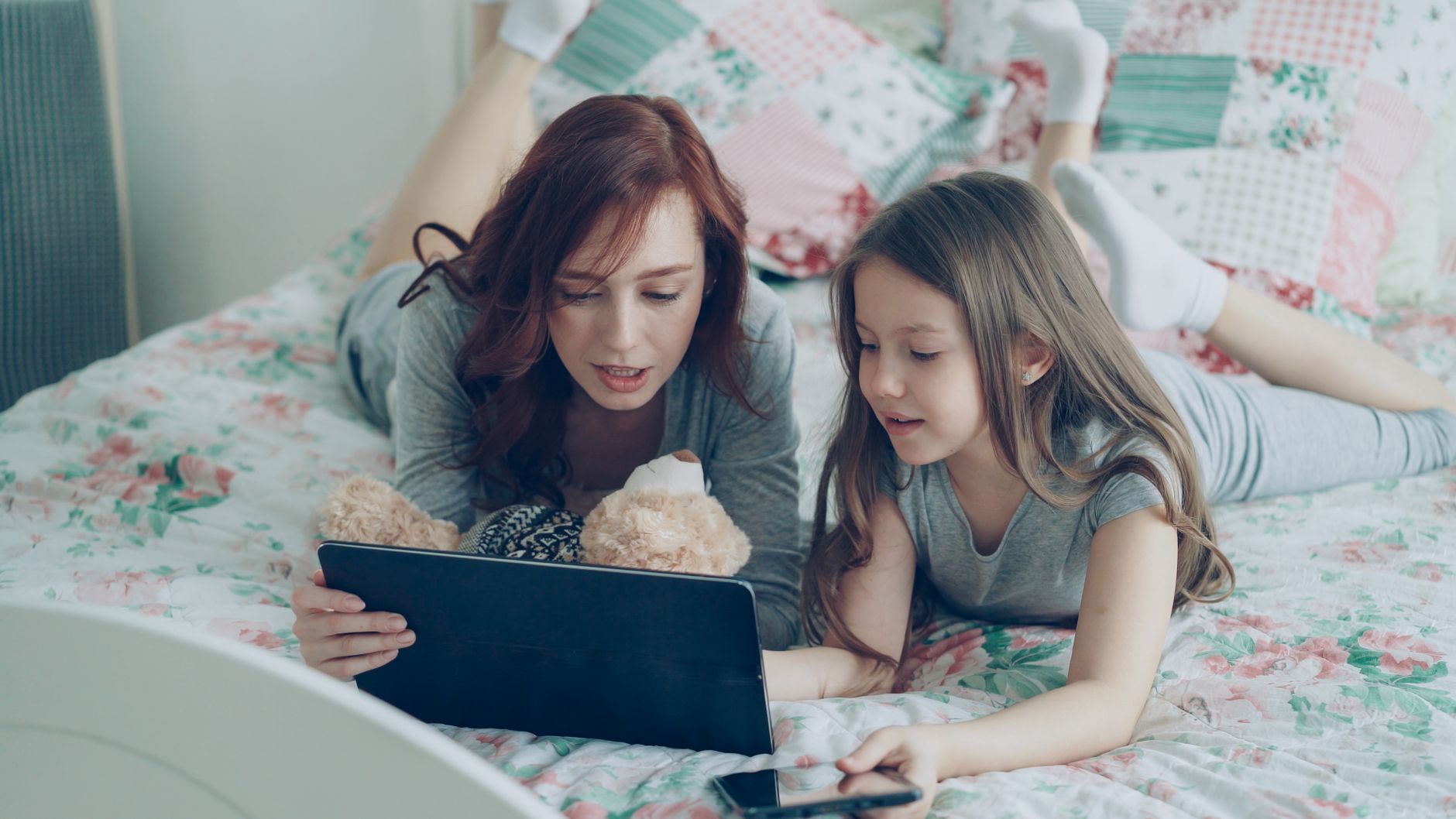 A mother and daughter enjoying time together using a tablet in a cozy bedroom setting.
