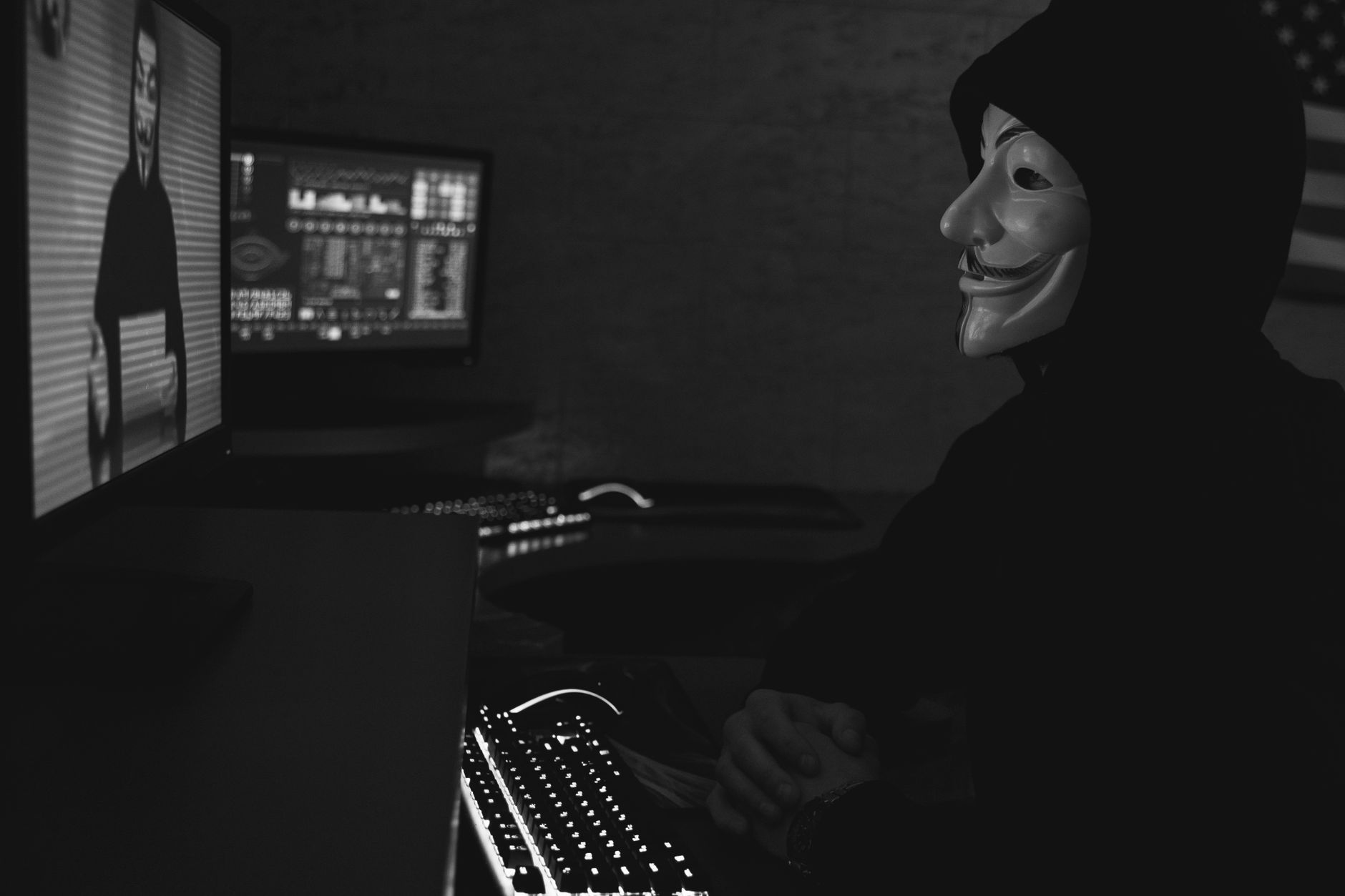Monochrome image of a hacker in a Guy Fawkes mask working on a computer in a dimly lit room.