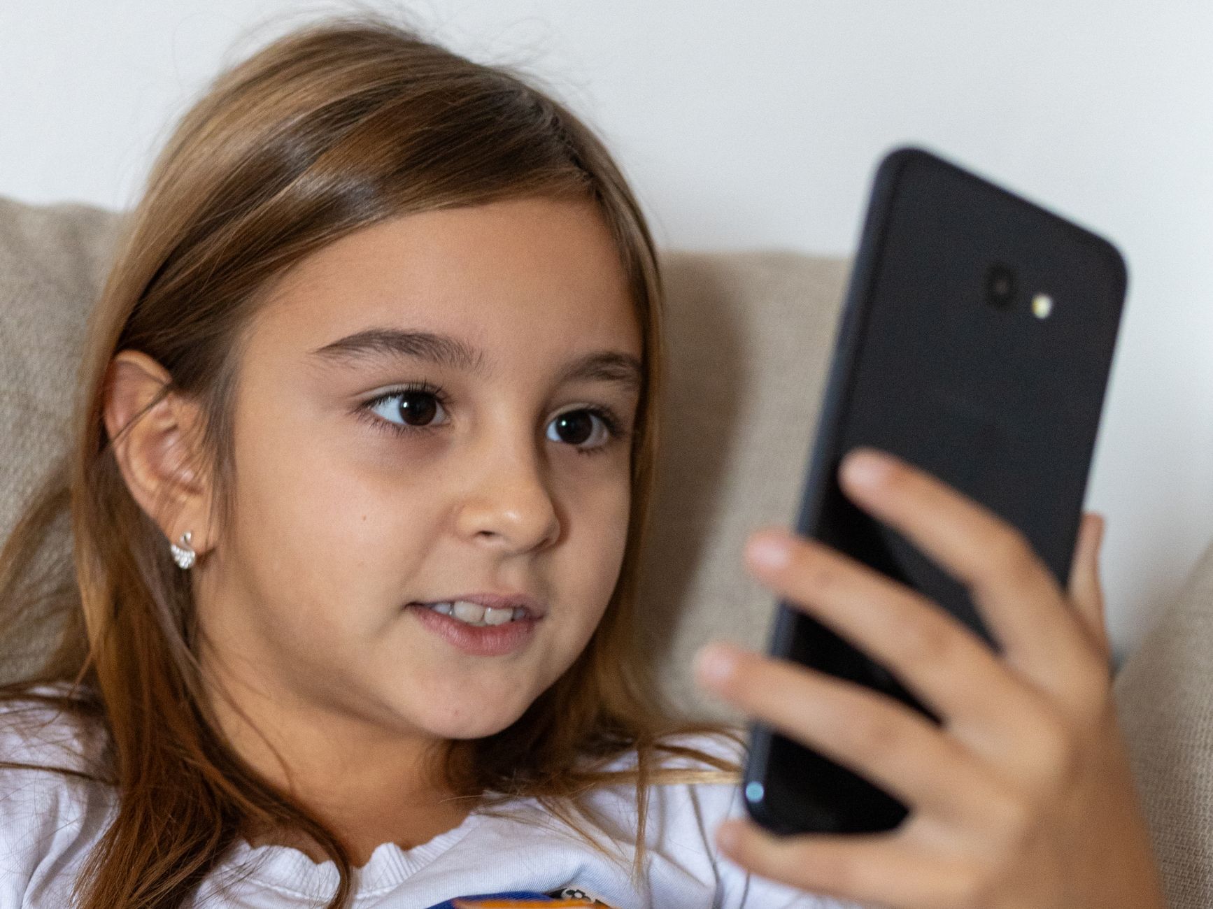 A young girl with long brown hair smiles while using a smartphone indoors.