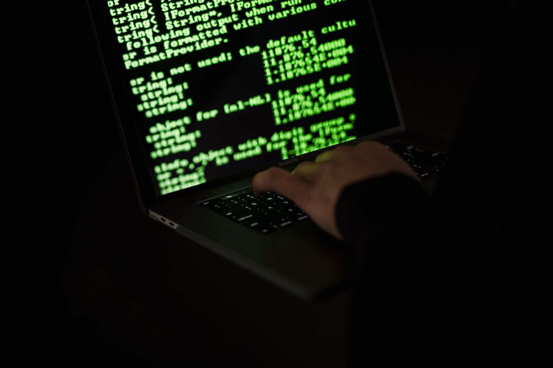Crop unrecognizable computer geek typing on netbook with codes on screen while hacking system in darkness