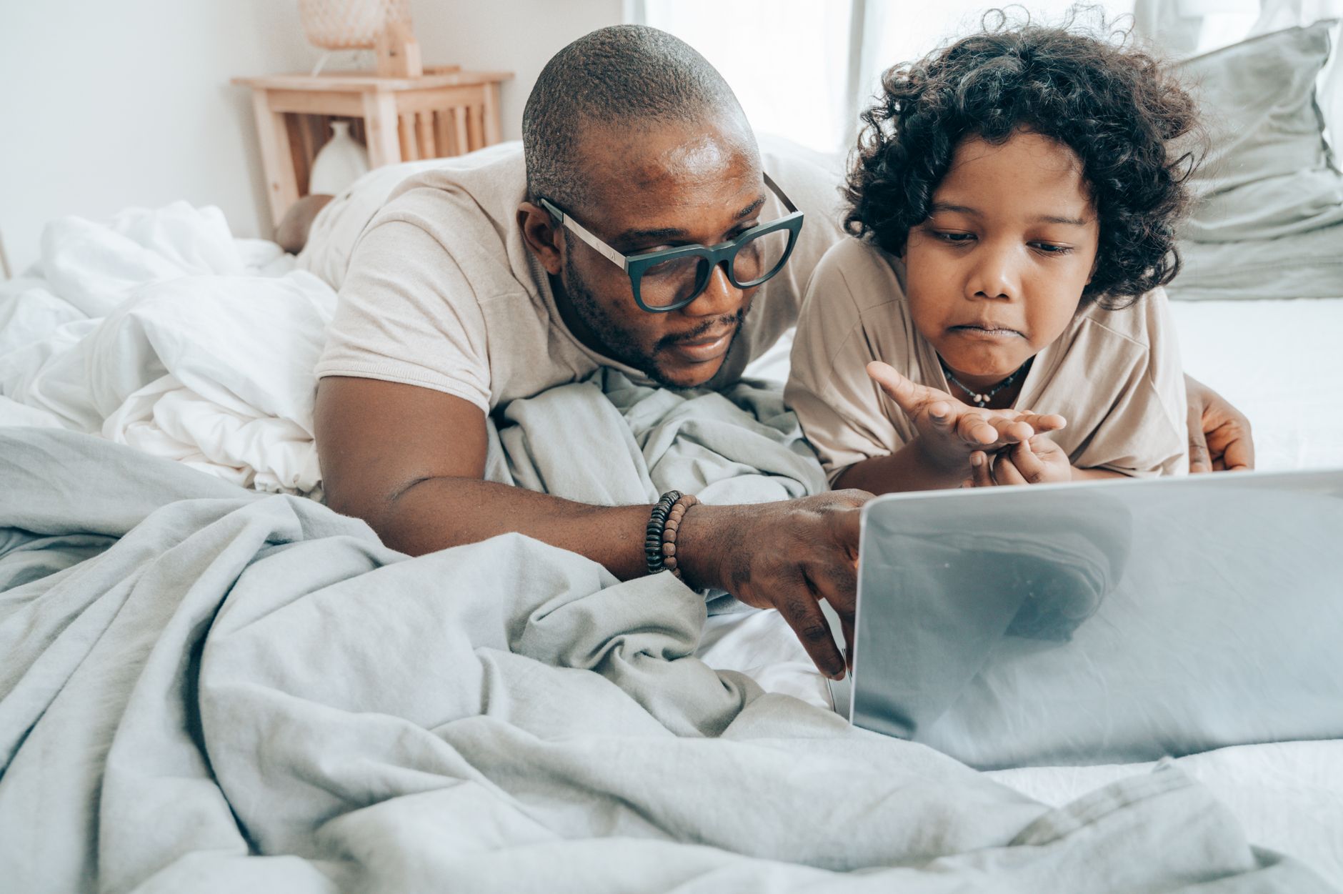 Focused African American male in glasses and child with curly hair in pajama lying on bed in bedroom and using laptop