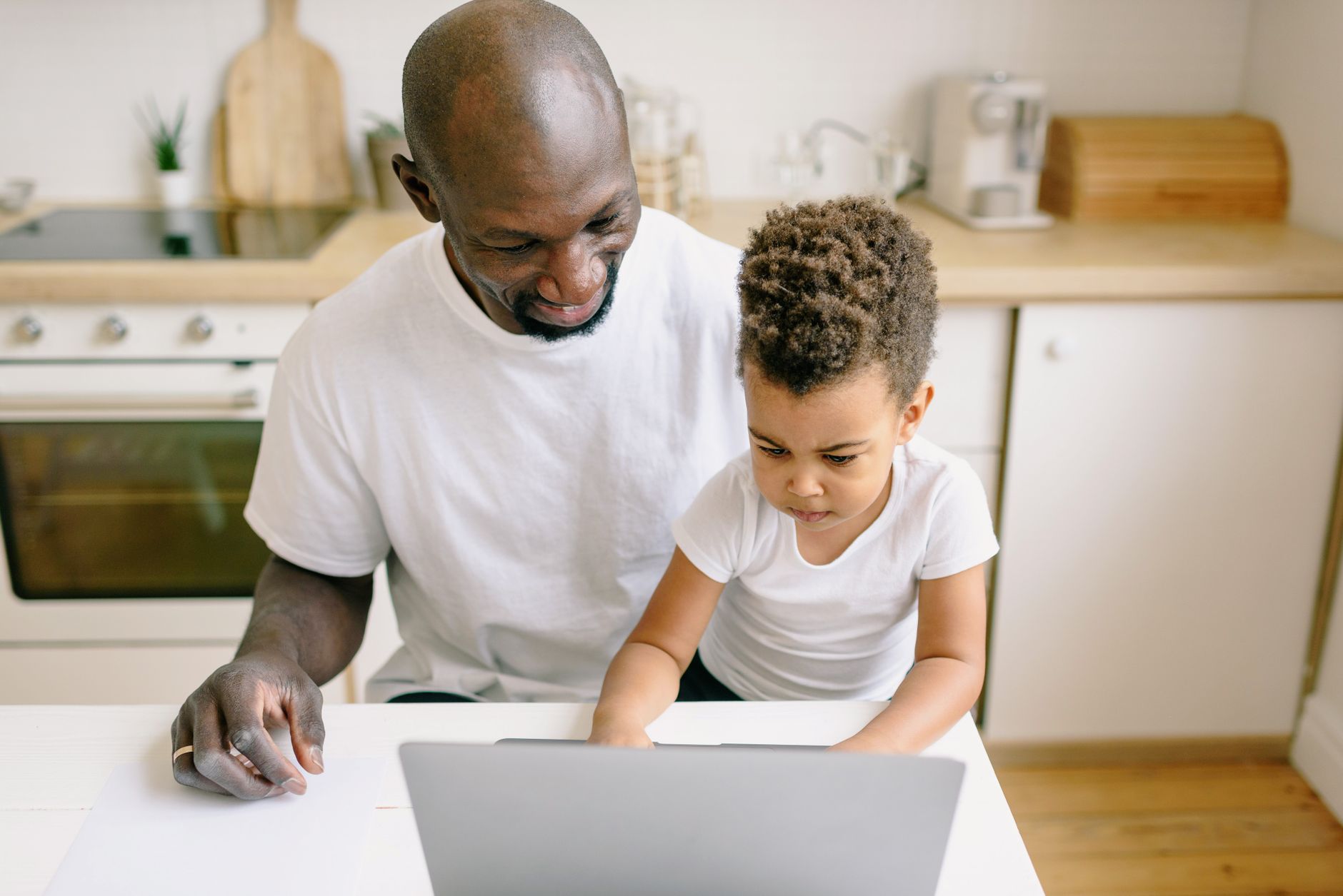 A father and son bonding while using a laptop together in a cozy kitchen setting.