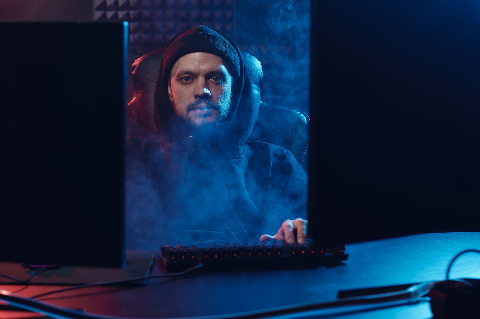 Dramatic scene of an adult man in a hoodie using a computer, surrounded by smoke in a dimly lit room.