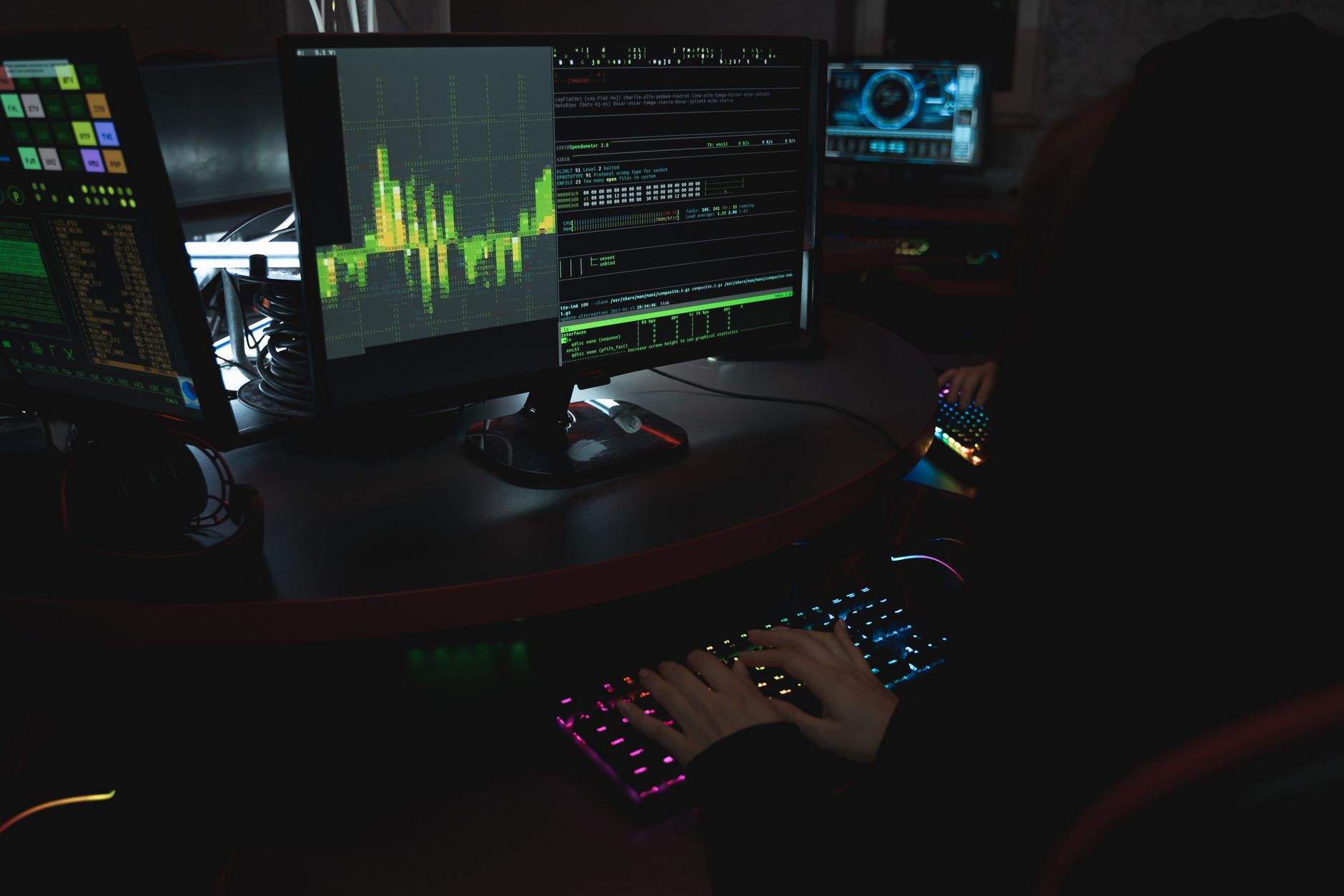 A cybersecurity expert in a dimly lit room is typing on a colorful keyboard with multiple screens displaying data.