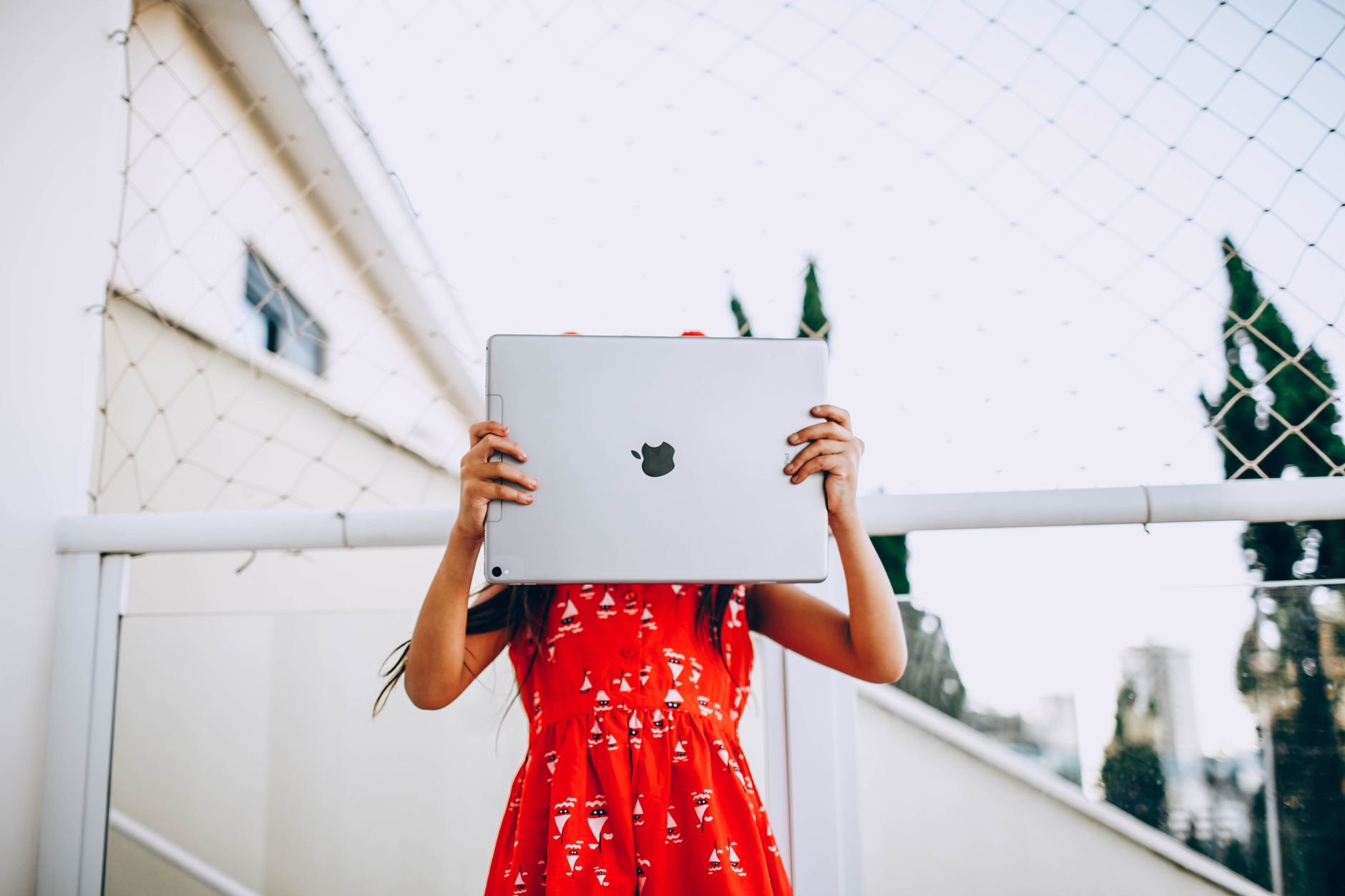 Low angle of unrecognizable child in dress standing on street and covering face with modern tablet