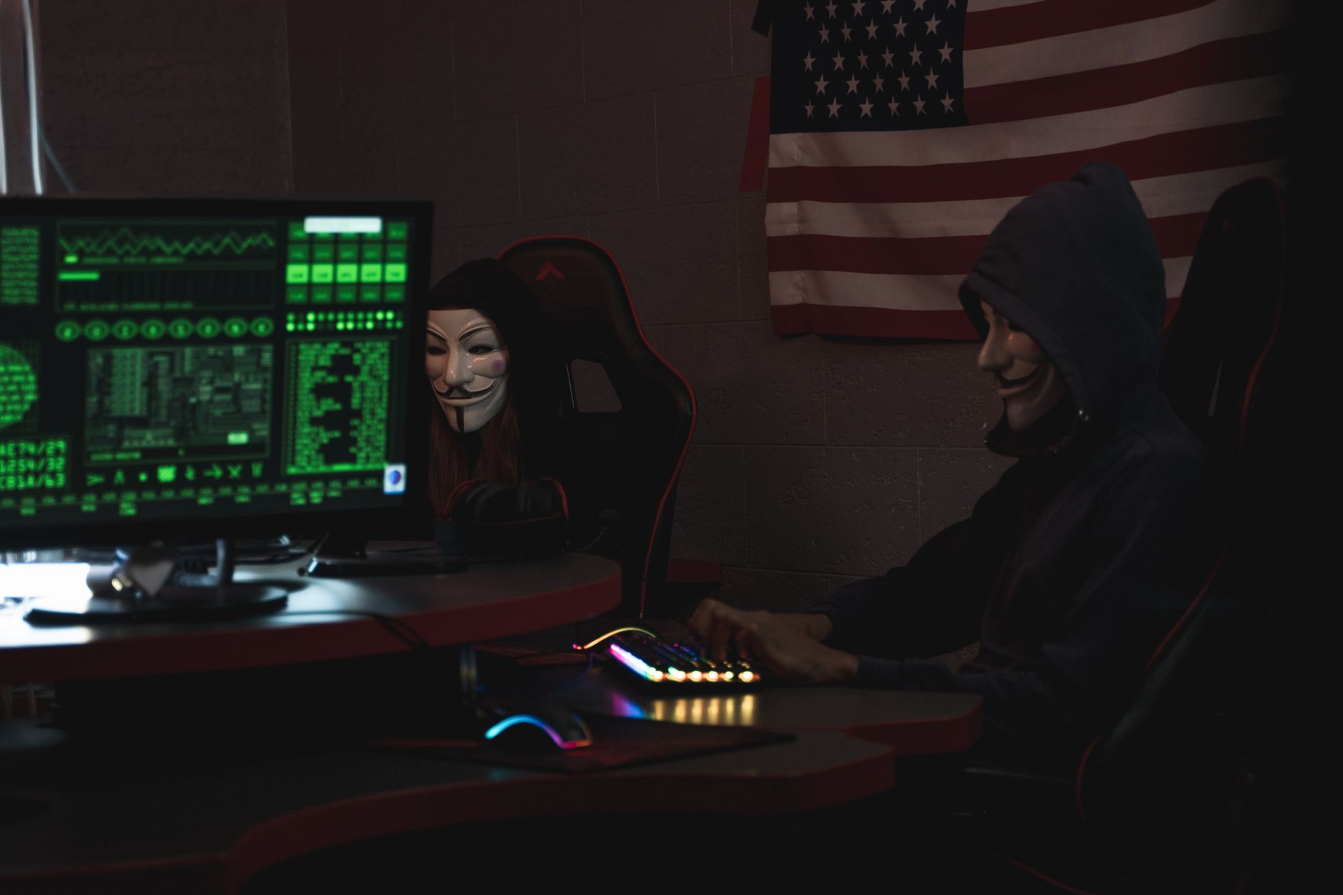 Two anonymous individuals wearing hacker masks use computers in a dark room with an American flag backdrop, highlighting cyber security them