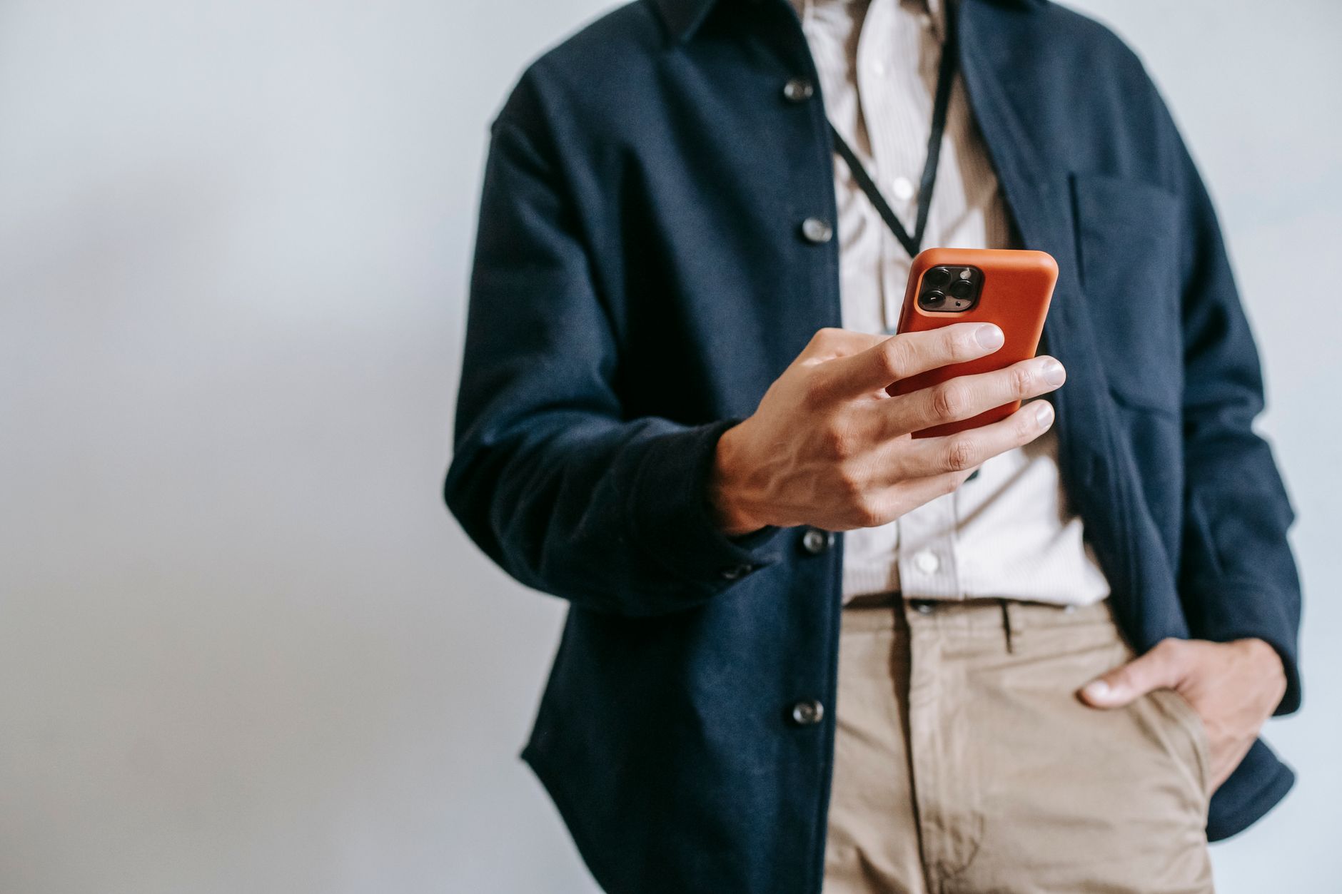 Low angle of crop unrecognizable male manager in trendy outfit and name badge messaging on mobile phone while standing near white wall with 