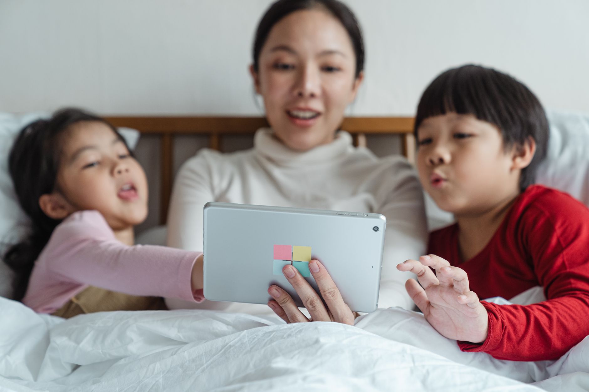 Interested Asian mother and little daughter and son in pajamas lying in comfortable bed choosing content in Internet on tablet while spendin