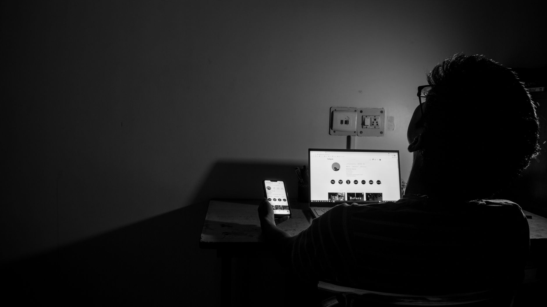 Silhouette of a man browsing the internet on a laptop and smartphone in a dark room.
